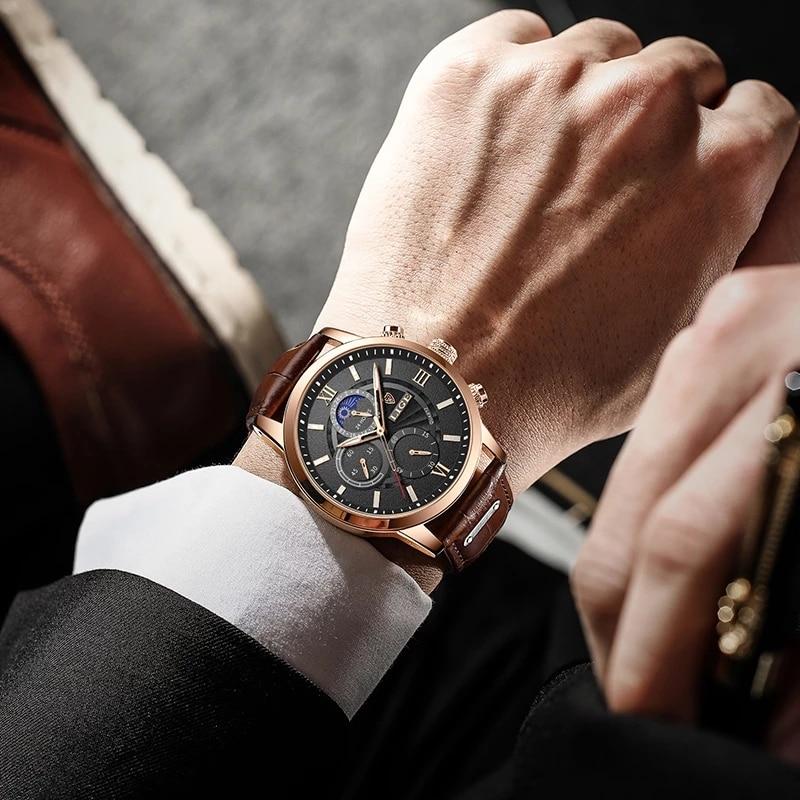 Aurelius Timepiece men's watch with gold case, black dial, and brown leather strap on a man's wrist, visible with a suit cuff.