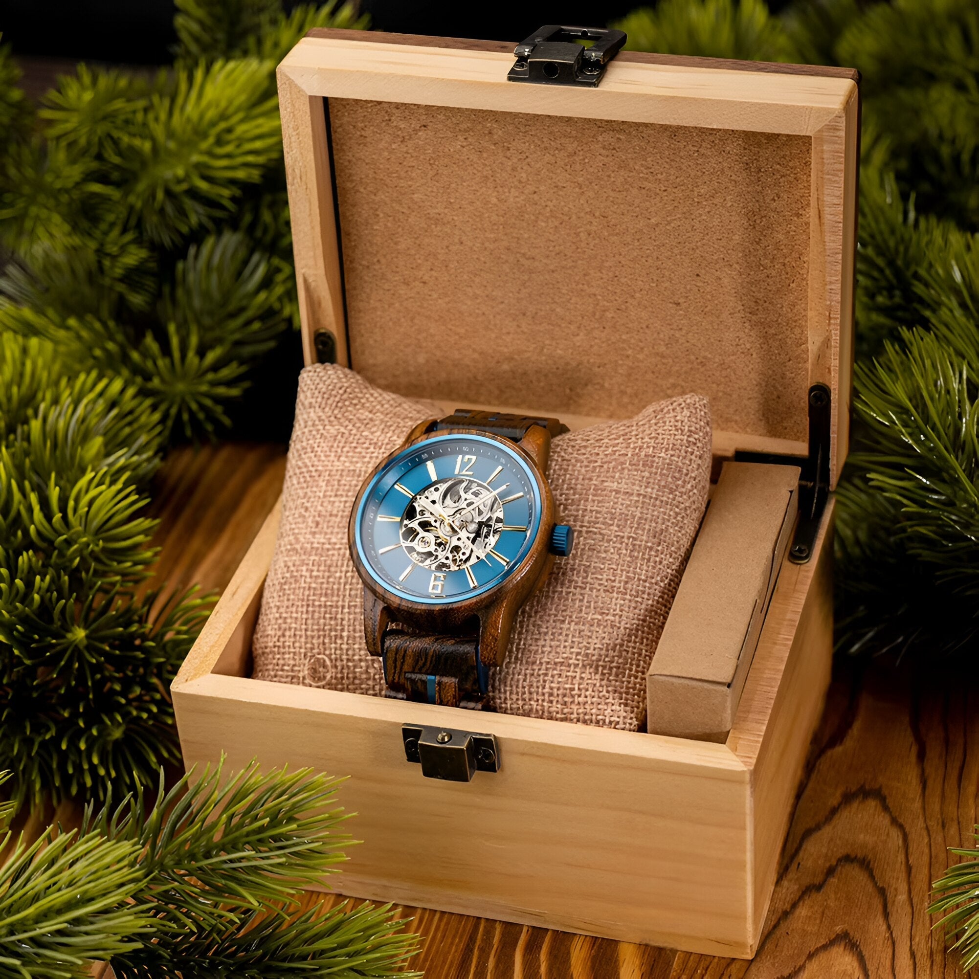 Cascadia Blue men's wooden watch with blue skeleton dial and stainless steel accents, displayed in an open light wood gift box with a burlap cushion, surrounded by pine needles.