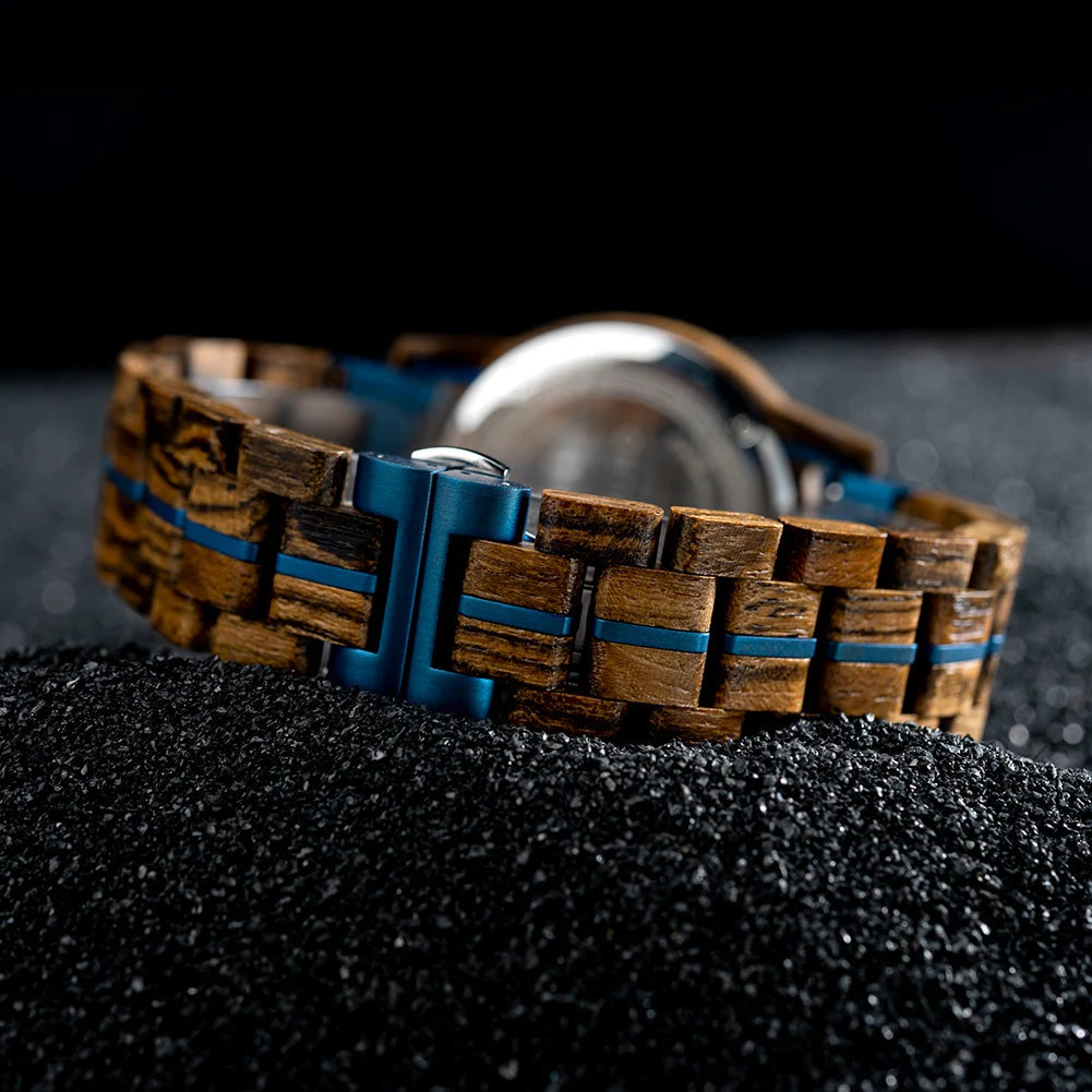 Close-up view of the Celestial Oak Men's Wooden Watch strap and blue metal clasp, showing blue accents on the wooden links, on a dark granular surface.