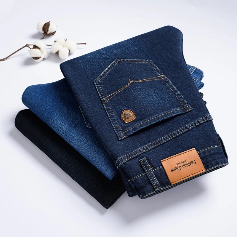 Stack of three folded Cityscape Trousers men's premium cotton classic fit pants in dark blue, light blue, and black, with a cotton plant branch in the background.
