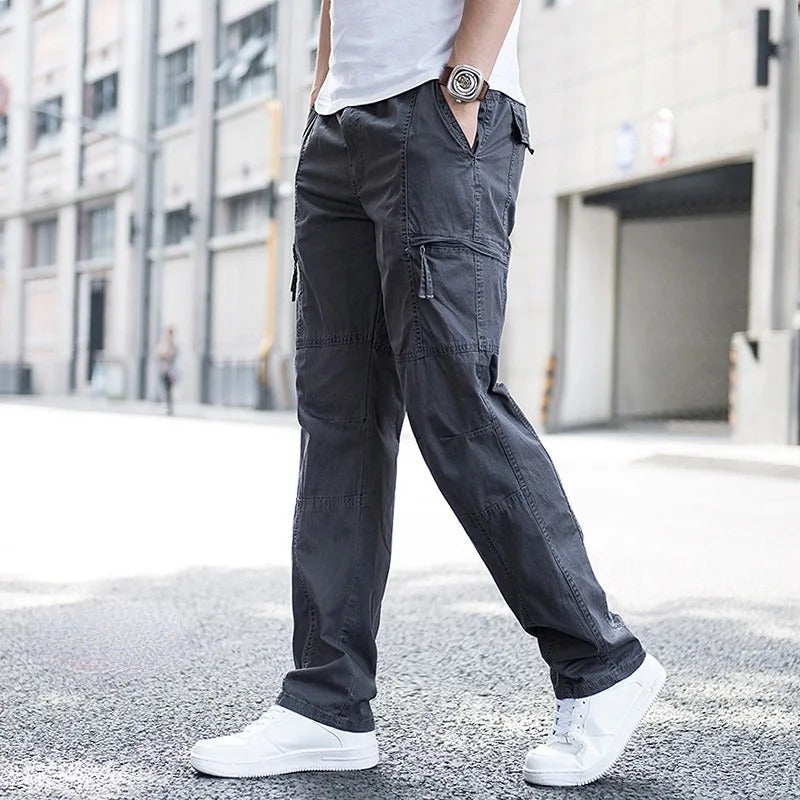 Male model showcasing Coastal Retreat dark gray cotton cargo pants with prominent utility pockets and white sneakers, walking on a city street. The pants offer a comfortable, relaxed fit suitable for outdoor clothing and everyday fashion.