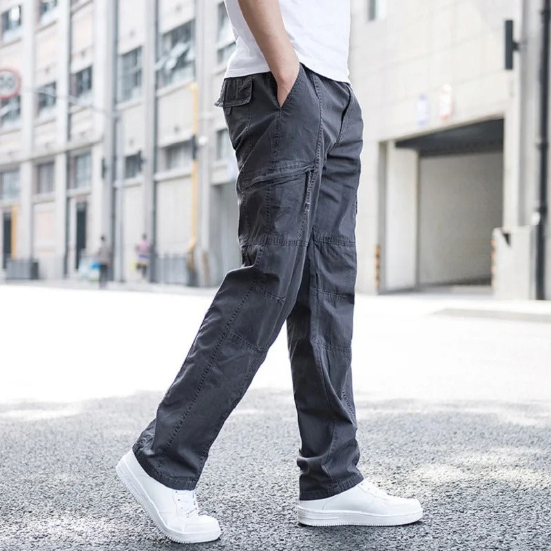 Male model wearing Coastal Retreat dark gray cotton cargo pants with utility pockets and white sneakers, walking on a city street. The pants feature a relaxed fit and durable fabric, perfect for casual wear.