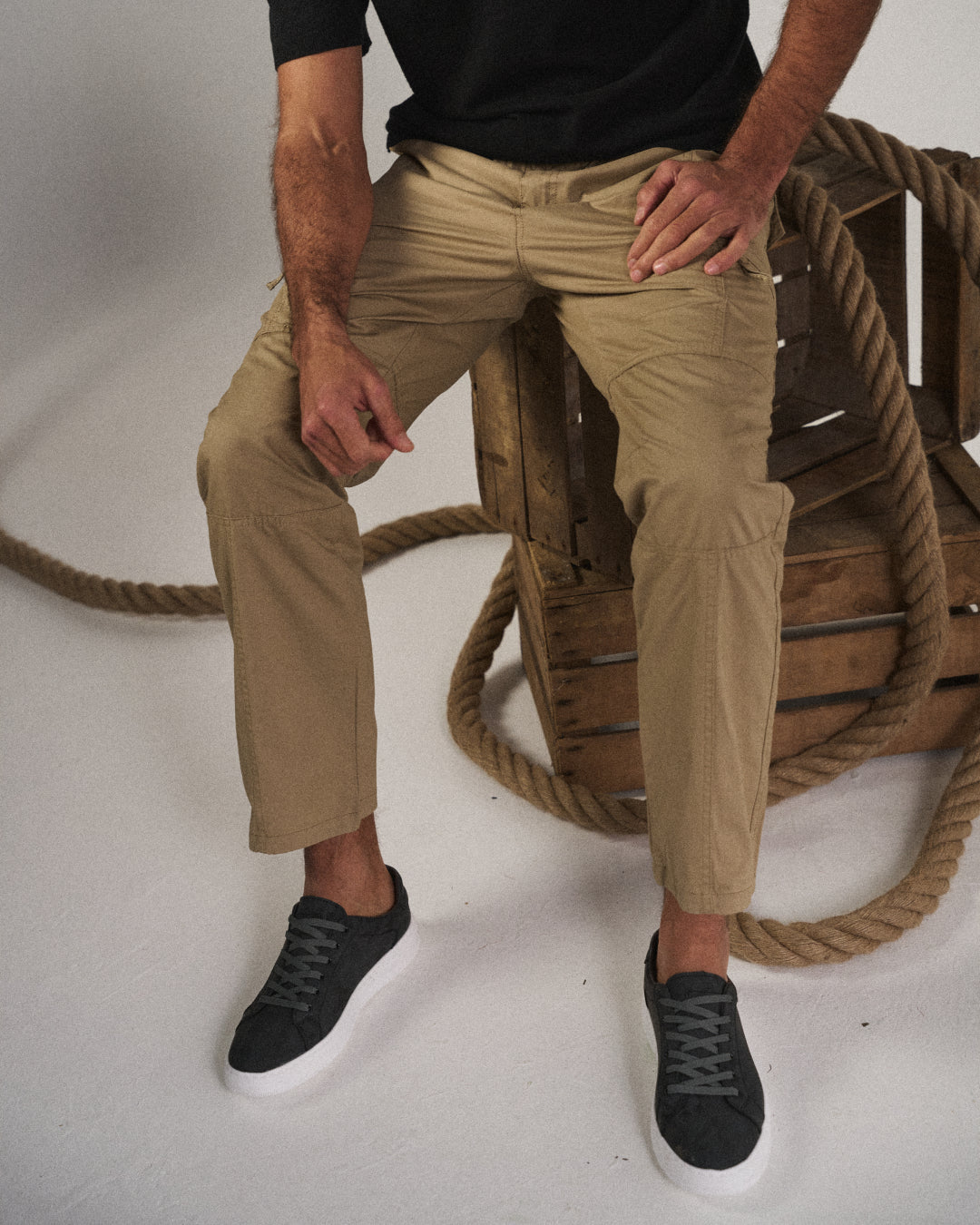 Close-up of a man sitting, showcasing the Khaki Coastal Retreat cotton cargo pants with utility pockets, paired with a black t-shirt and black sneakers, with wooden crates and rope in the background.