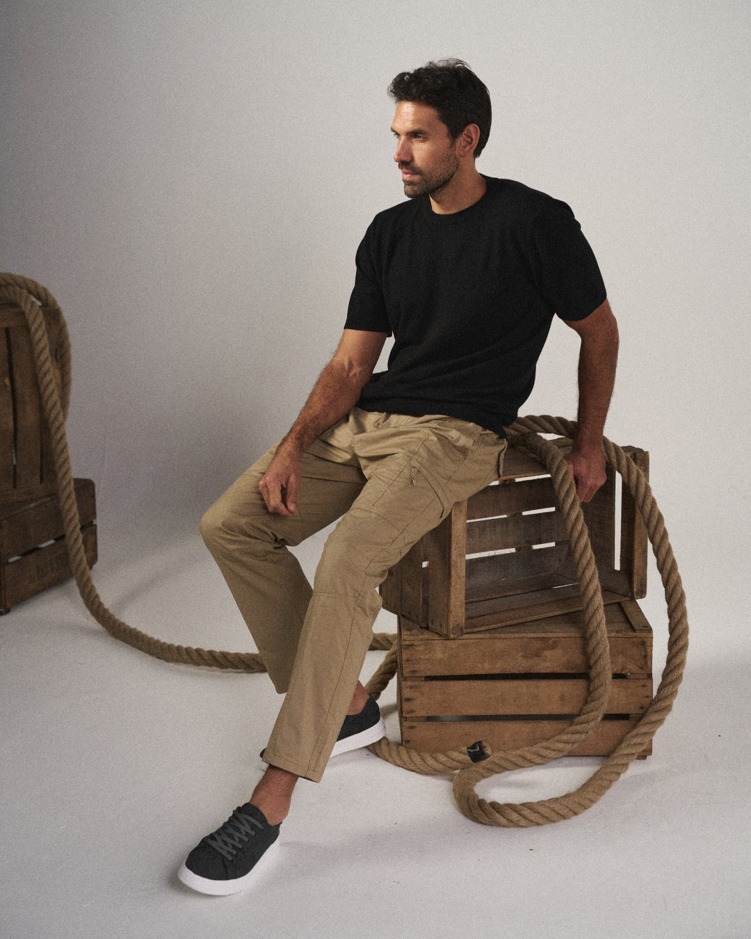 A man wearing khaki Coastal Retreat cargo pants with utility pockets and a black t-shirt, sitting casually on wooden crates with thick ropes in a studio setting.