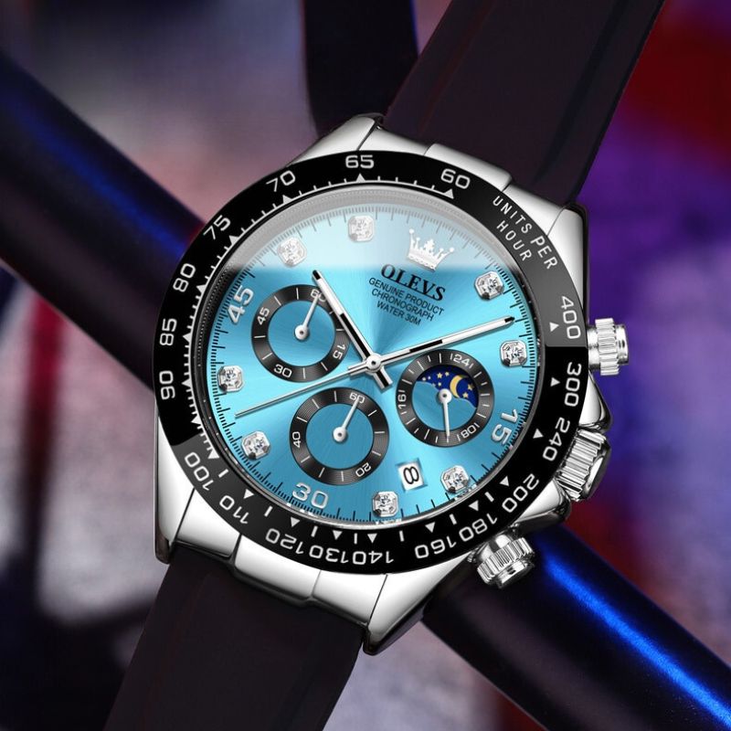 A detailed close-up of the Elysium Timepiece men's watch featuring a blue dial, silver case, black silicone strap, and chronograph subdials, resting on a dark reflective surface.