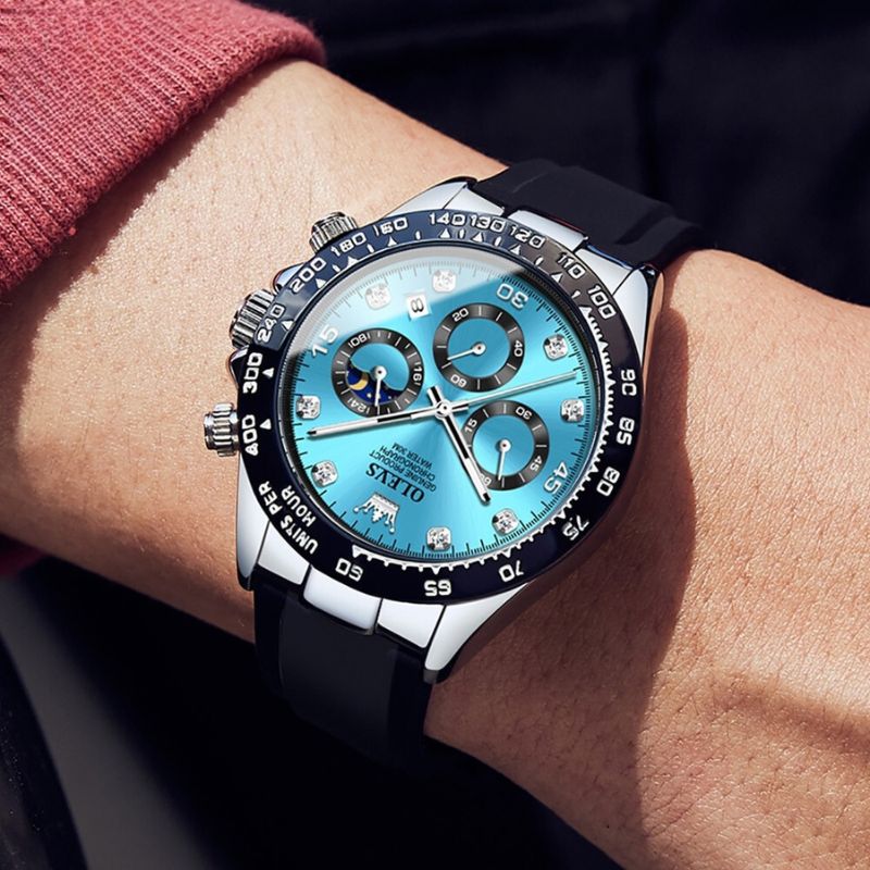 A close-up shot of a man's wrist wearing an Elysium Timepiece men's watch with a blue dial, silver case, and black silicone strap, against a blurred red background.