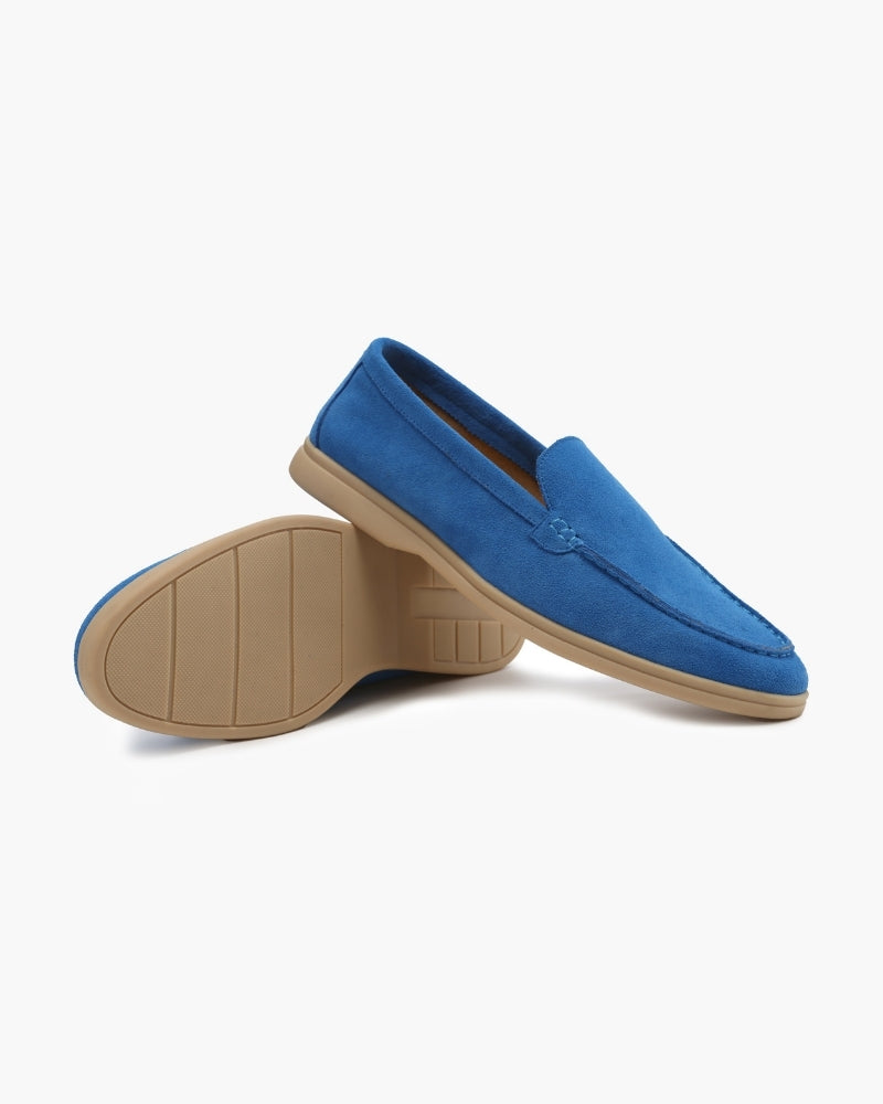 Heritage Luxe men's blue suede loafers with classic stitching, one shoe upright the other inverted to display textured beige sole, on a white background.