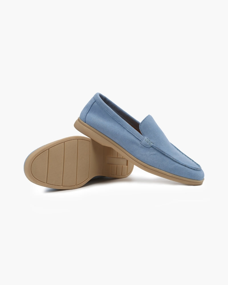 A pair of light blue suede men's loafers, one standing upright showing the side and top, the other on its side revealing the tan sole with a grip pattern, against a white background.