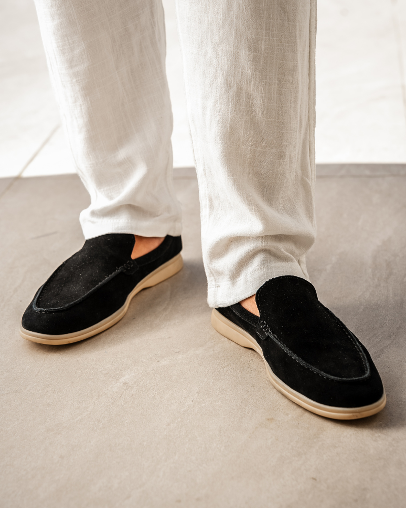 A man's feet wearing Heritage Luxe black suede loafers with classic stitching and a tan sole, paired with white linen pants, standing on a light grey tiled floor.
