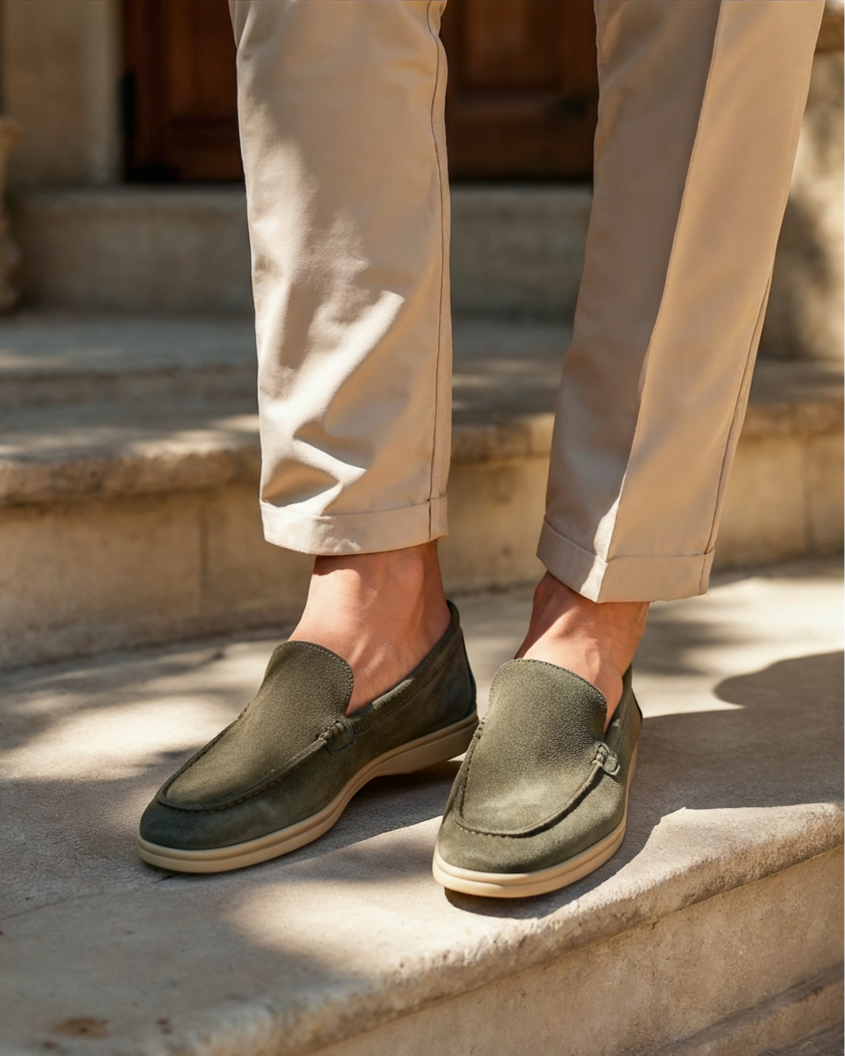 Men's Heritage Luxe olive green suede loafers with classic stitching, worn by a person in beige trousers, standing on outdoor steps.