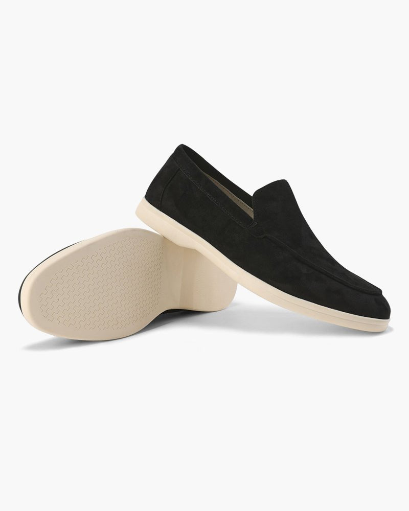 A pair of Heritage Luxe black suede loafers, one upright showing the side profile and classic stitching, the other on its side revealing the textured sole pattern.