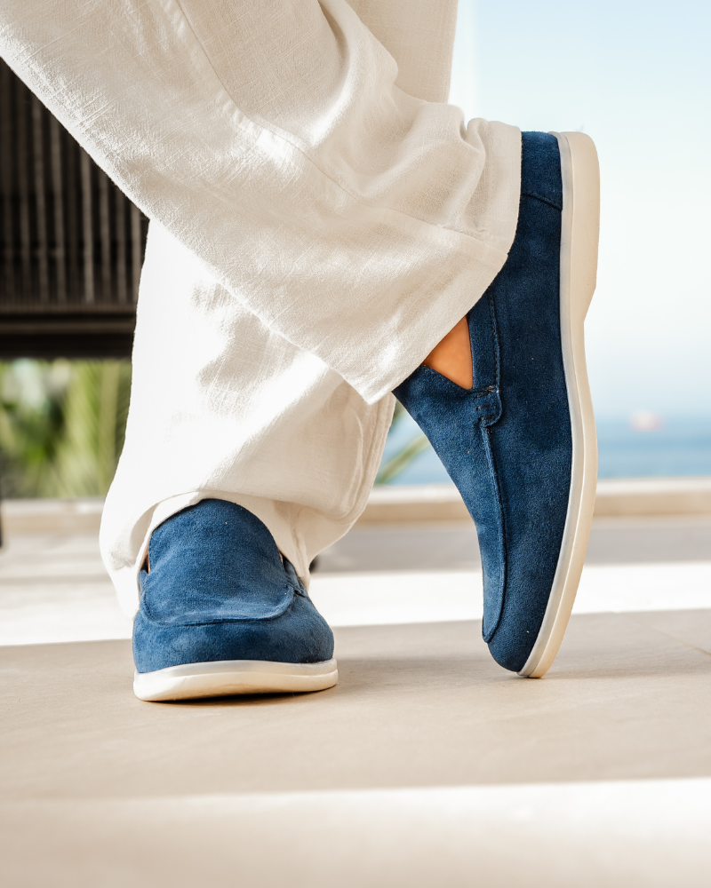 Close-up of a man's feet wearing Heritage Luxe Marine Blue suede loafers with cream soles and white pants, standing on a tiled patio with a blurred ocean view in the background. Showcases fashionable men's footwear.