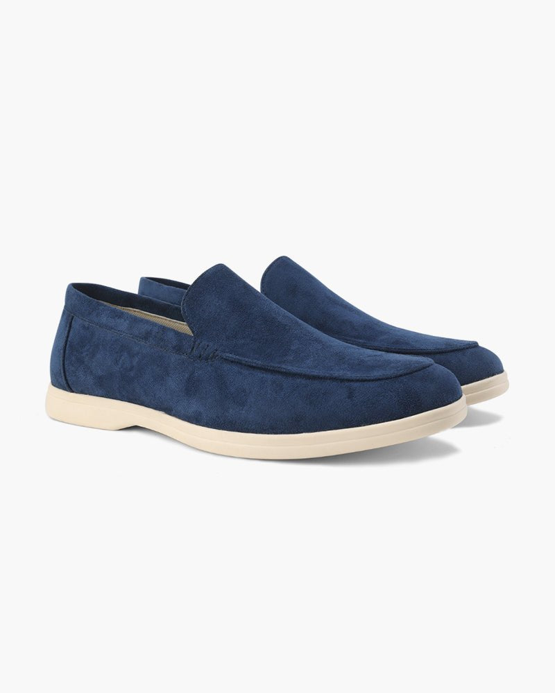 A pair of Heritage Luxe Marine Blue suede loafers with classic stitching, featuring a light cream sole, presented on a clean white background. These men's stylish slip-ons offer luxury and comfort.