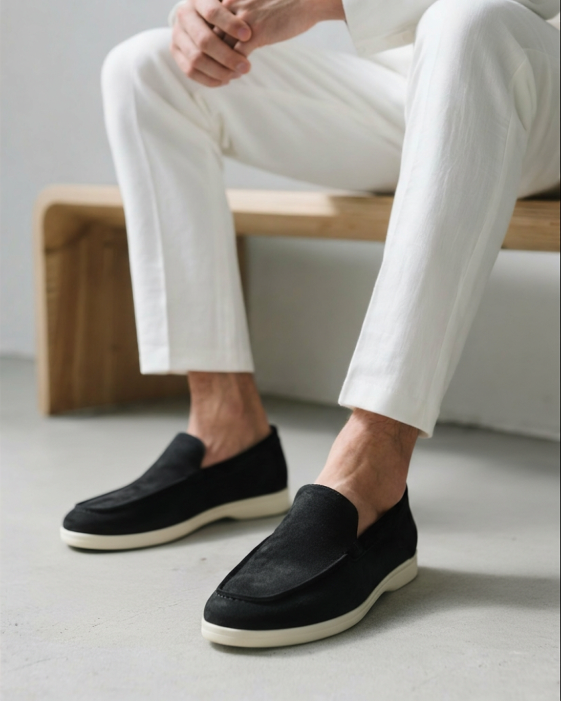 Close-up of a man wearing black Heritage Luxe suede loafers with classic stitching and white soles, paired with white pants, sitting on a wooden bench.