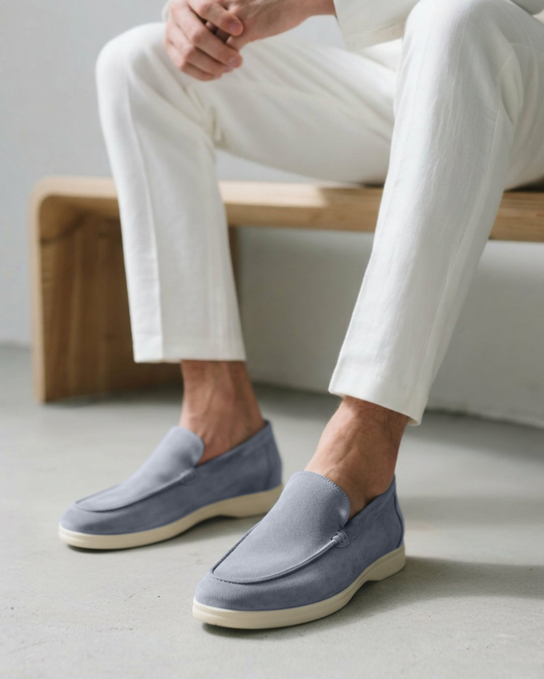 Close-up of a man wearing fog blue Heritage Luxe suede loafers with classic stitching and white soles, paired with white pants, sitting on a wooden bench.