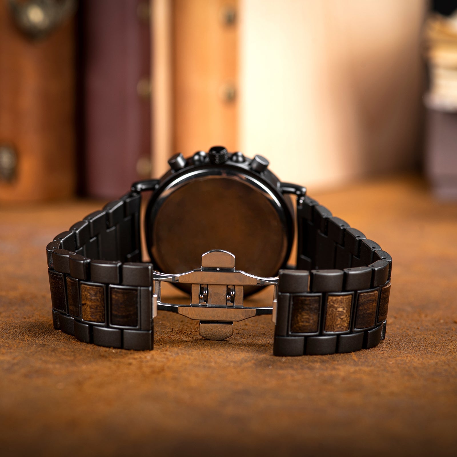 Back view of the Heritage Oak men's watch, showcasing the metal case back and the butterfly clasp on the natural wood and dark metal link strap, resting on a textured brown surface.