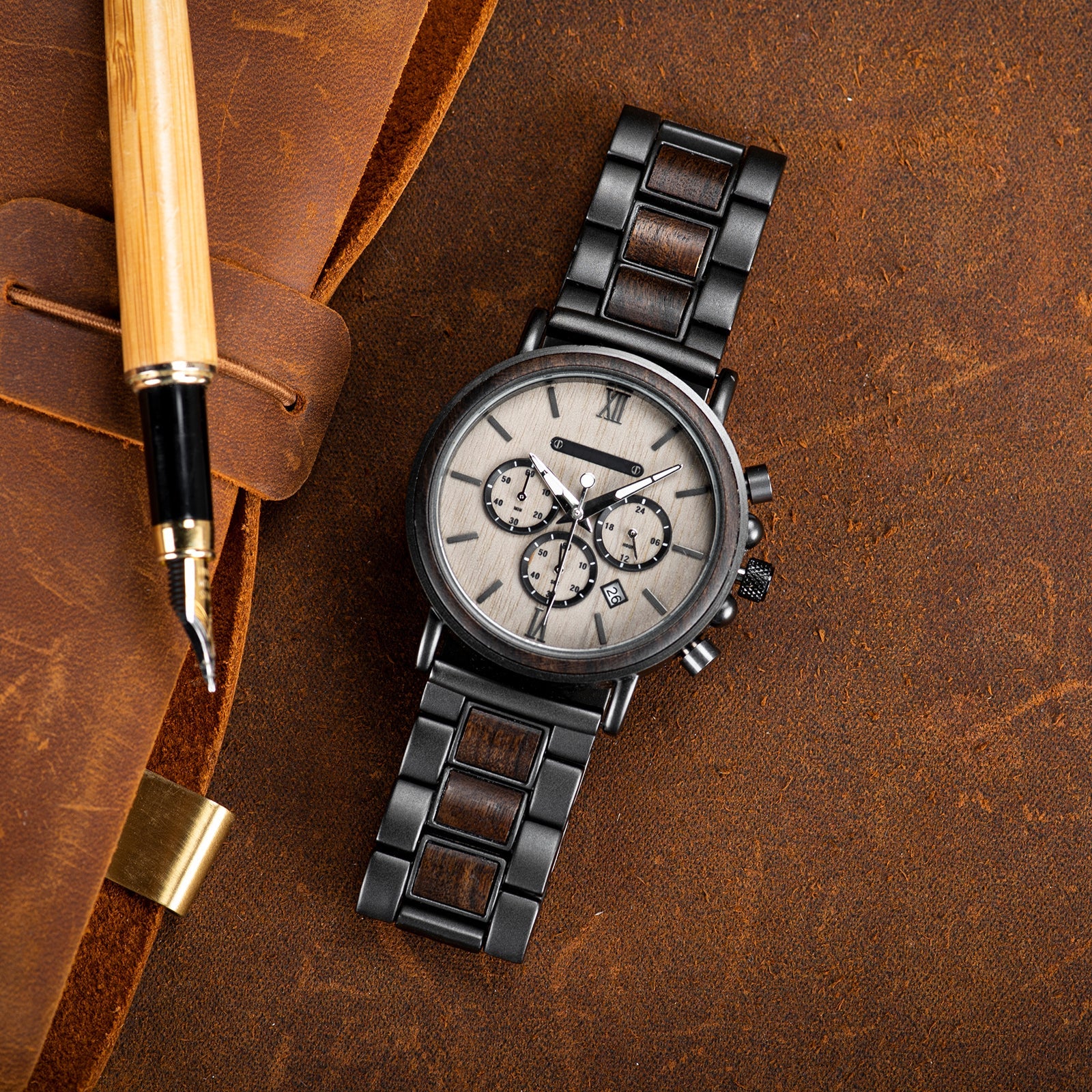 Heritage Oak men's watch with natural wood and dark metal strap, light dial, and chronograph features, displayed on a brown leather surface next to a bamboo pen.