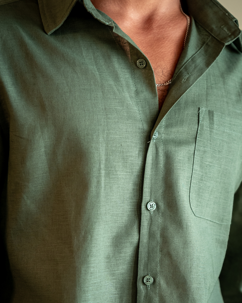 Close-up detail of a dark green Linen Essence men's shirt, highlighting the natural texture of the 100% linen fabric, the front buttons, and the chest pocket. A silver chain is visible around the wearer's neck.