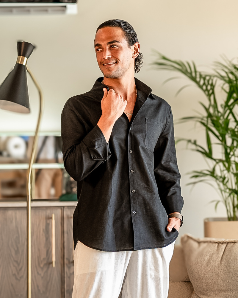 A smiling man wearing a black Linen Essence classic fit shirt and white linen pants, standing indoors next to a lamp and a plant, looking to the side.