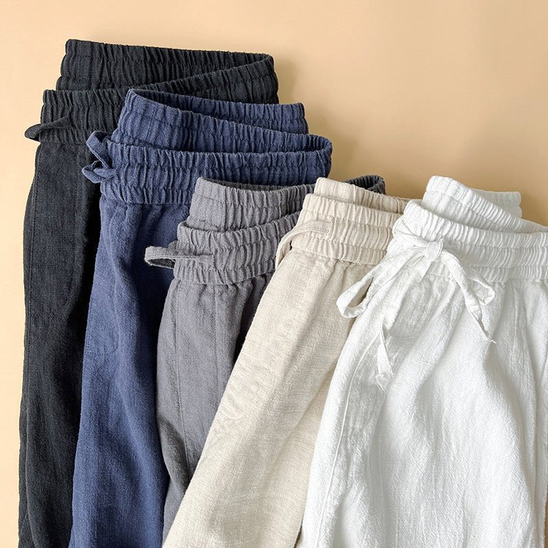 Five pairs of Luxe Retreat Comfy Linen Pants with Relaxed Fit, stacked and folded, showcasing the elastic waistbands and drawstrings in black, blue, gray, khaki, and white colors.