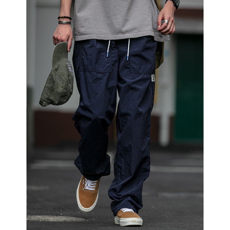A man models Sakura Breeze waterproof, breathable, relaxed fit casual pants in navy, paired with a grey t-shirt and holding a green cap, standing outdoors.