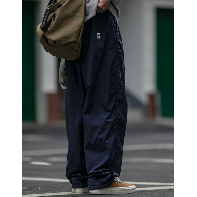 Side view of a man modeling Sakura Breeze waterproof, breathable, relaxed fit casual pants in navy, paired with a grey t-shirt and shoulder bag, showing a logo detail.
