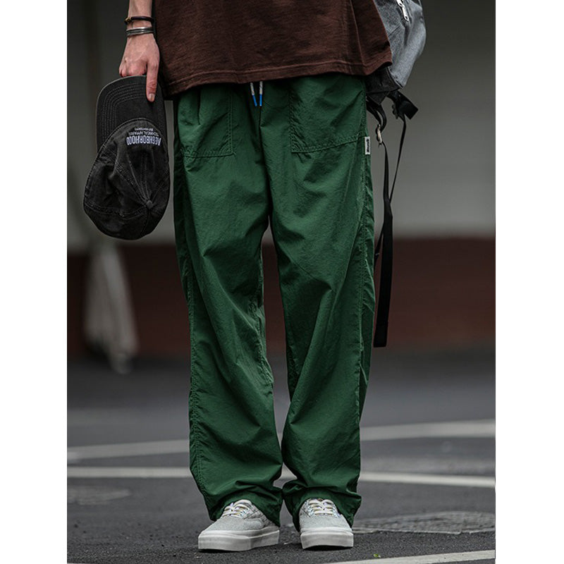 Men's Sakura Breeze waterproof breathable casual pants in green, shown on a model wearing white sneakers and a brown t-shirt, holding a black cap. Emphasizes the relaxed fit and casual style.