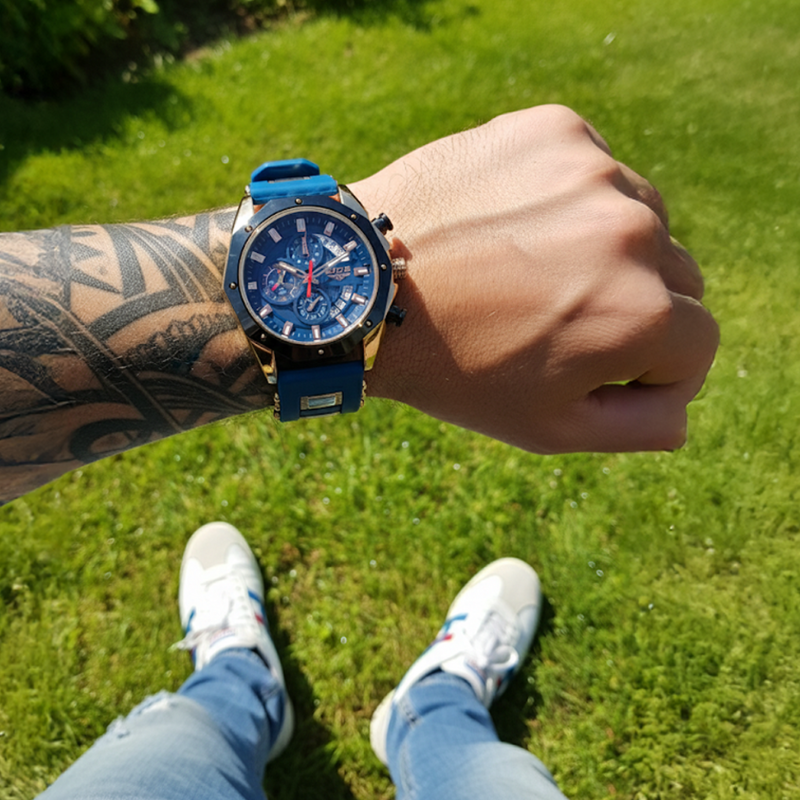 Overhead view of a man wearing the Skyline Aviator men's watch with a blue dial and gold case on his tattooed arm, standing on green grass with white sneakers and blue jeans.