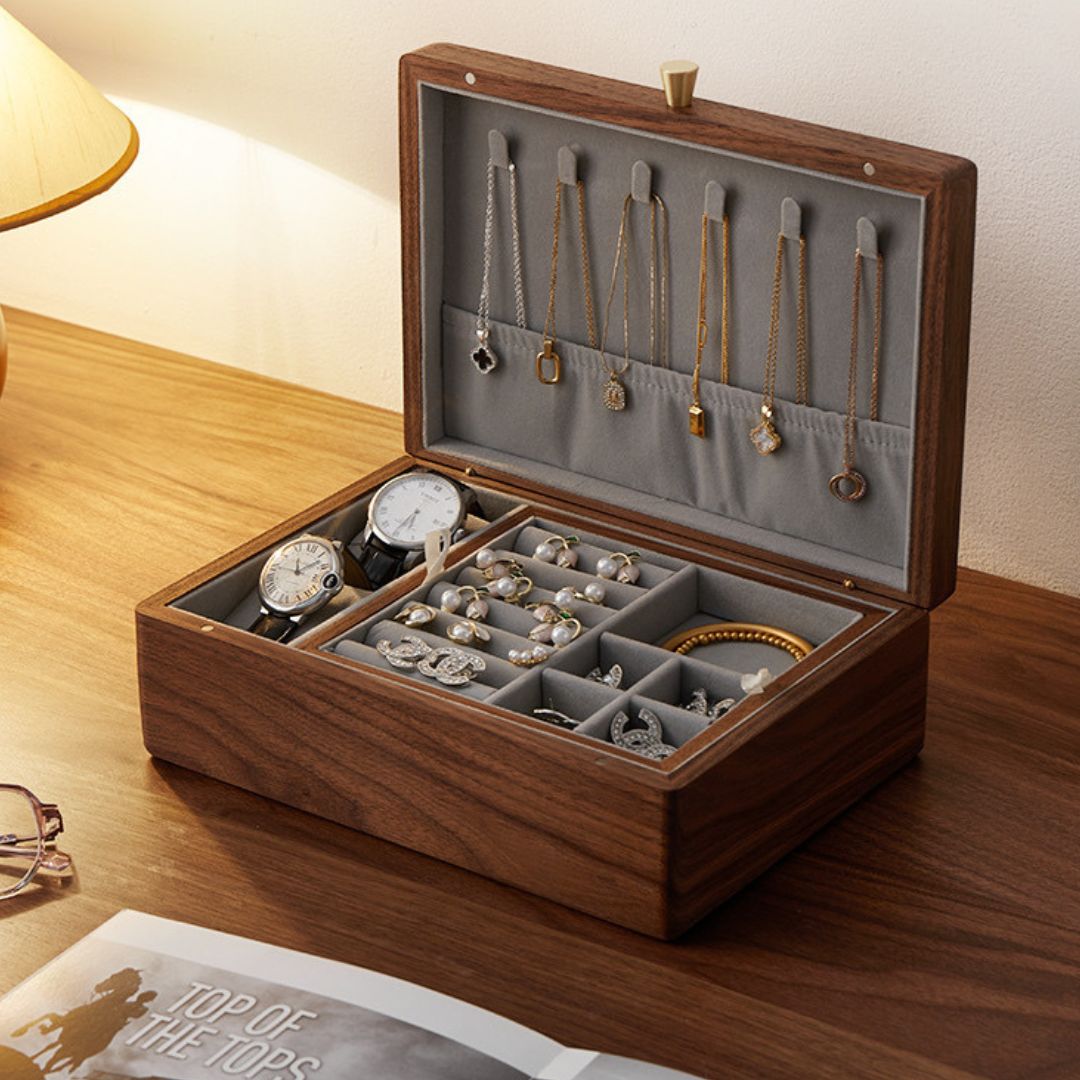 An open Timber Haven solid walnut wood jewelry box with a removable tray, displaying two watches, various rings, earrings, and necklaces. The box sits on a wooden desk next to a lamp and an open magazine.