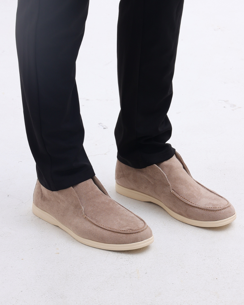 Mocha brown suede loafers with classic detailing worn by a person in black pants, shown from a standing view.