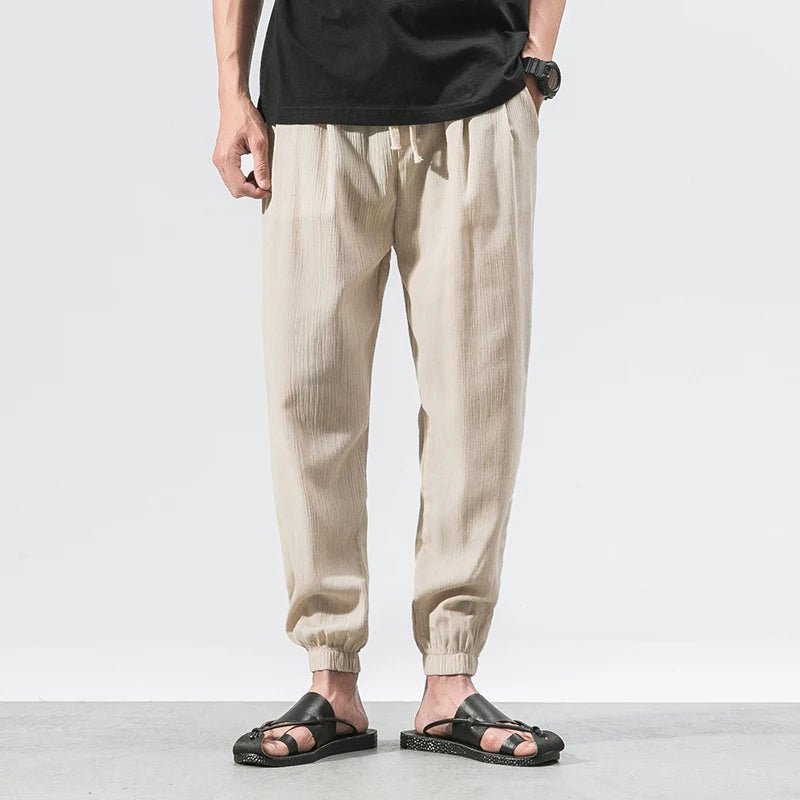 Full shot of a man wearing beige Wanderer's Path boho style linen pants with a relaxed fit, paired with a black t-shirt and black sandals. The pants feature a drawstring waist and elastic cuffs, showcasing their comfortable and casual design.