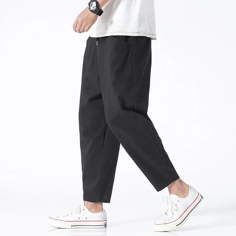 A man models Zenith Cropped Trousers in black, made from a cotton-linen blend, paired with white sneakers. The lightweight, tailored pants feature a drawstring waist and side pockets, ideal for smart-casual summer attire.