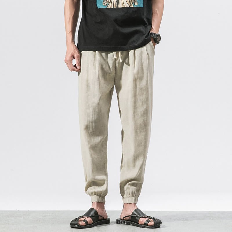 Full body shot of a man wearing Zenith Stones Beige linen drawstring pants with elastic cuffs, paired with a black t-shirt and black sandals. The pants feature side pockets and a comfortable, relaxed fit.