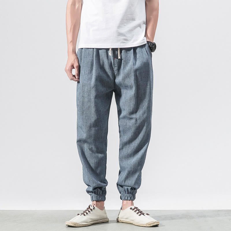 Full body shot of a man wearing Zenith Stones SteelBlue linen drawstring pants with elastic cuffs, paired with a white t-shirt and light-colored casual shoes. The pants feature side pockets and a relaxed fit.