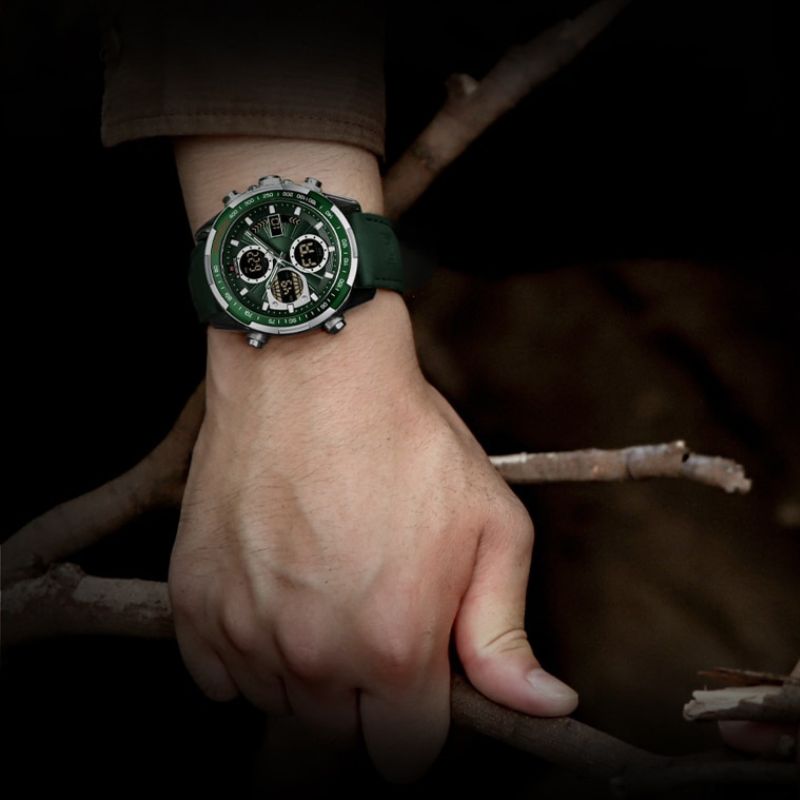 A close-up of a man's wrist wearing an Aether Voyage men's quartz watch with a silver case, green dial, and green leather band, holding tree branches in a dimly lit setting. The watch features luminous hands and a chronograph display.