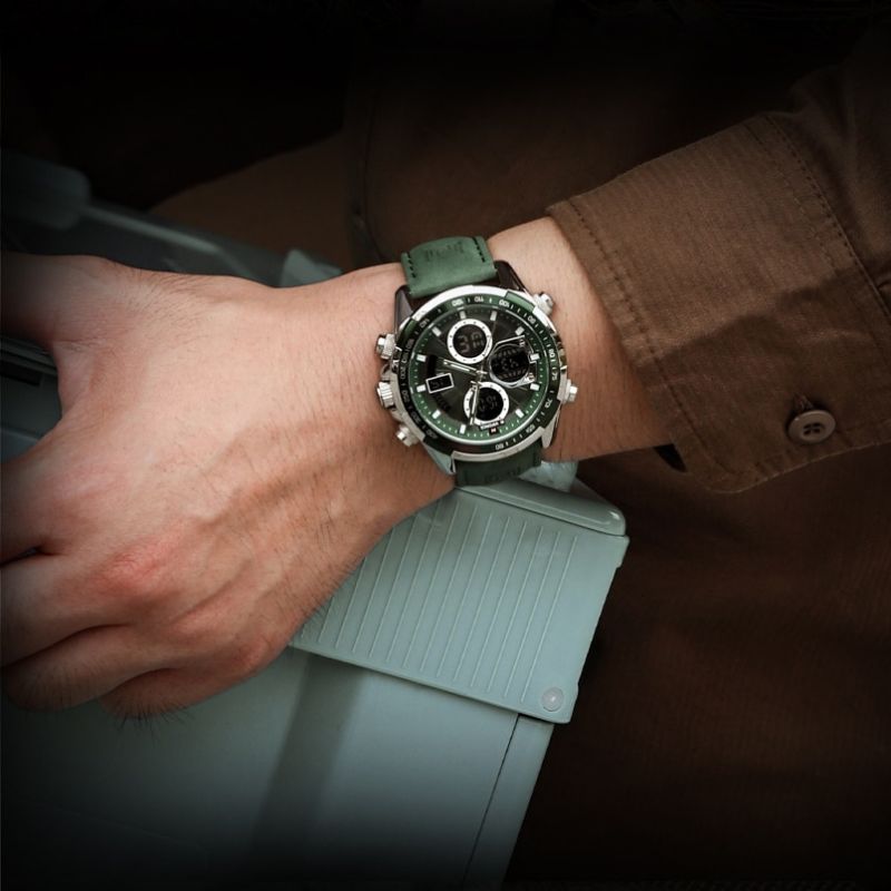 A man's wrist wearing an Aether Voyage men's quartz watch with a silver case, green dial, and green leather band, resting his hand on a light gray object. The watch displays luminous hands and chronograph sub-dials.