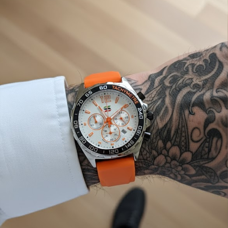Apex Dusk men's quartz watch with luminous hands and date display, silver case, white dial, and orange silicone strap, worn on a man's tattooed arm with a white shirt cuff.