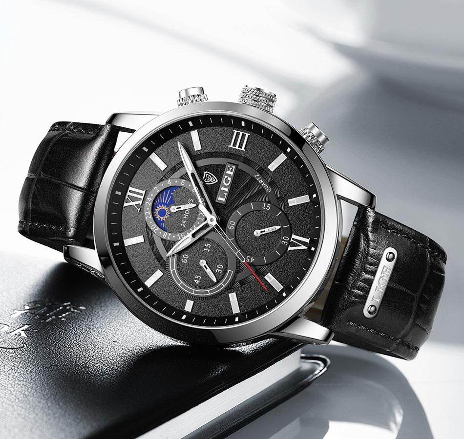 A close-up view of the Aurelius Timepiece men's watch featuring a black leather strap, a silver-tone case, and a black dial with Roman numerals, three functional subdials, and a moon phase indicator. The watch hands are luminous.