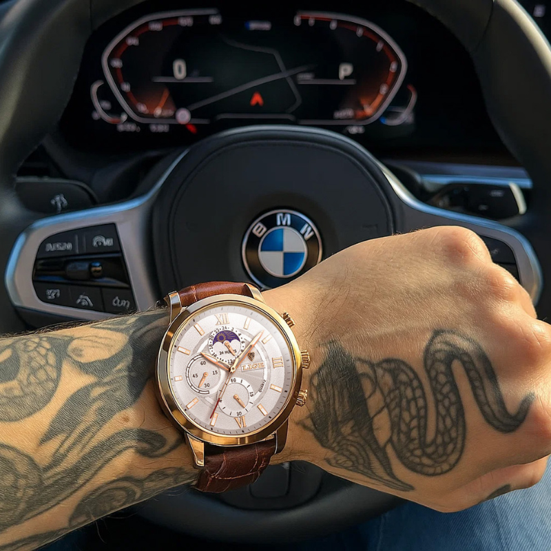Aurelius Timepiece men's watch with gold case, white dial, and brown leather strap on a man's tattooed arm holding a car steering wheel.
