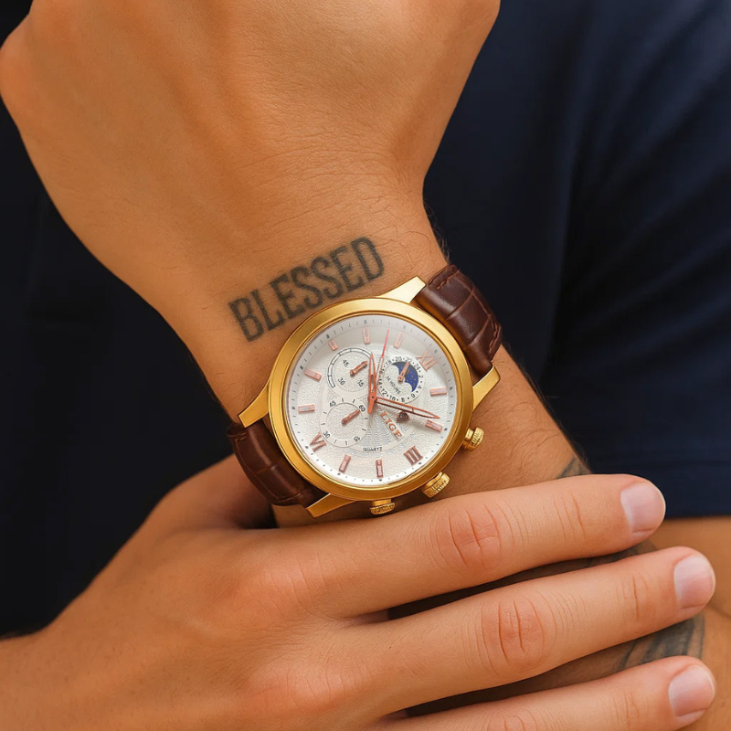Aurelius Timepiece men's watch with gold case, white dial, and brown leather strap on a man's wrist with a 'Blessed' tattoo.