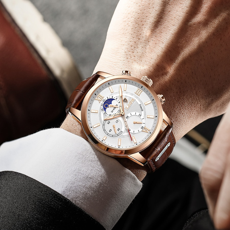 A close-up of a man's wrist wearing the Aurelius Timepiece, a gold-cased men's watch with a white dial, brown leather strap, luminous hands, and moon phase feature. The watch features multiple subdials and Roman numerals.