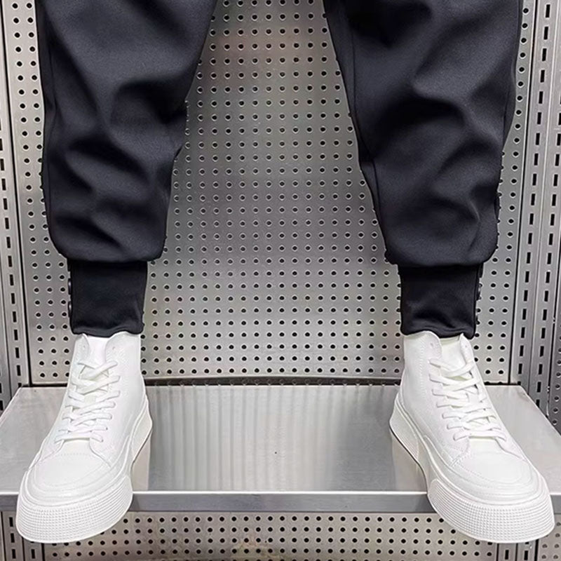 Black pleated harem trousers with elasticated cuffs worn with white sneakers, close-up view of lower legs on a metal display.