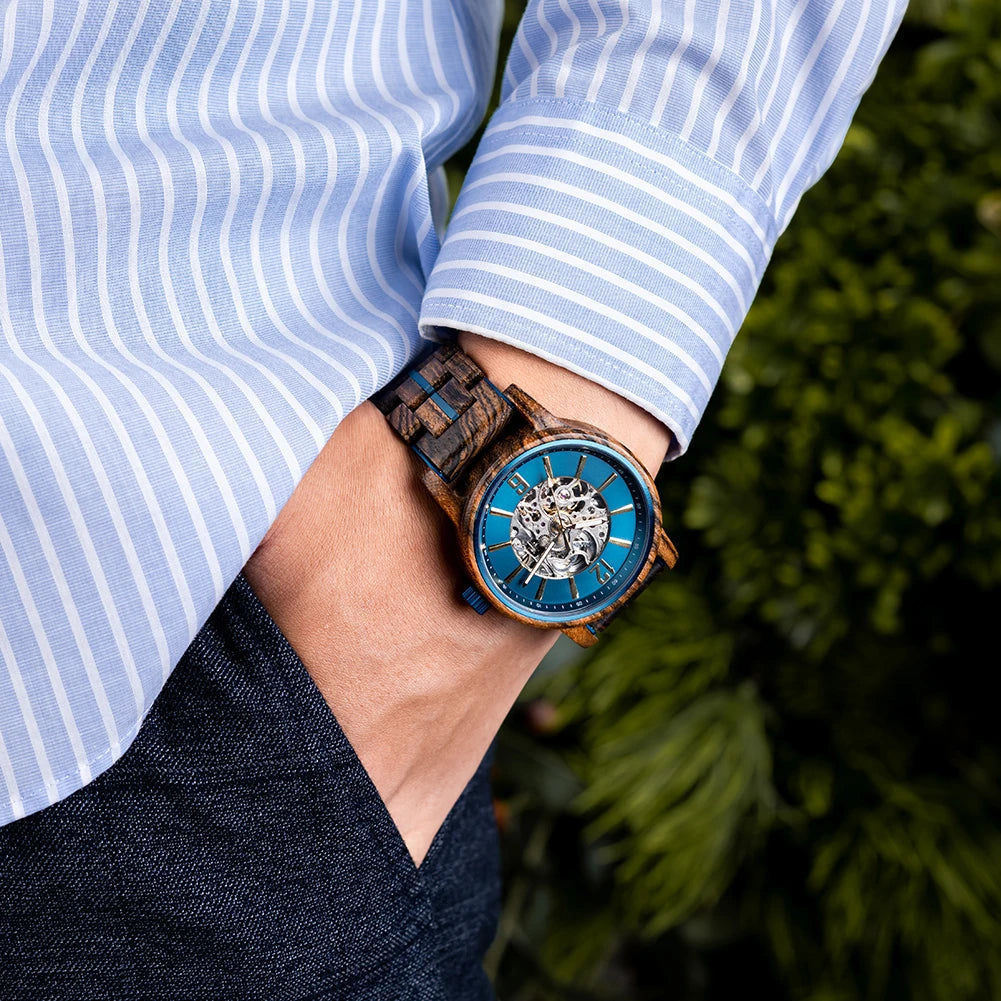 Cascadia Blue men's wooden watch with stainless steel accents worn on a man's wrist, paired with a light blue striped shirt and dark pants, set against a blurred green outdoor background.