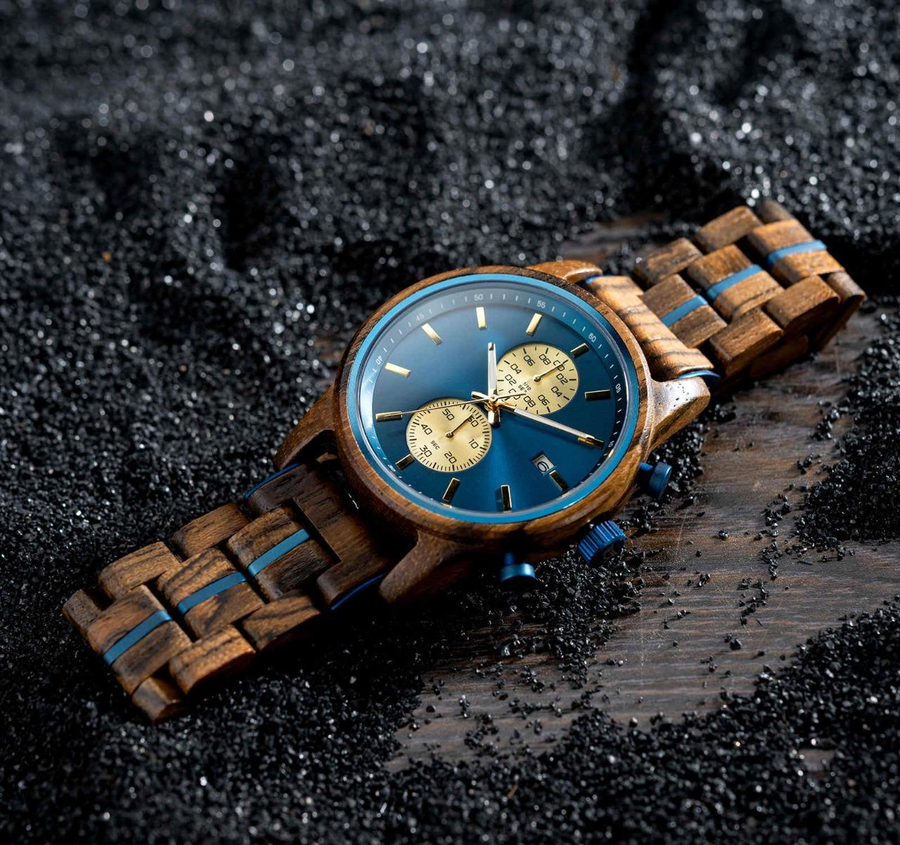 A close-up shot of the Celestial Oak men's wooden watch featuring a blue dial with gold subdials and a wooden strap with blue accents, resting on a dark, textured surface.