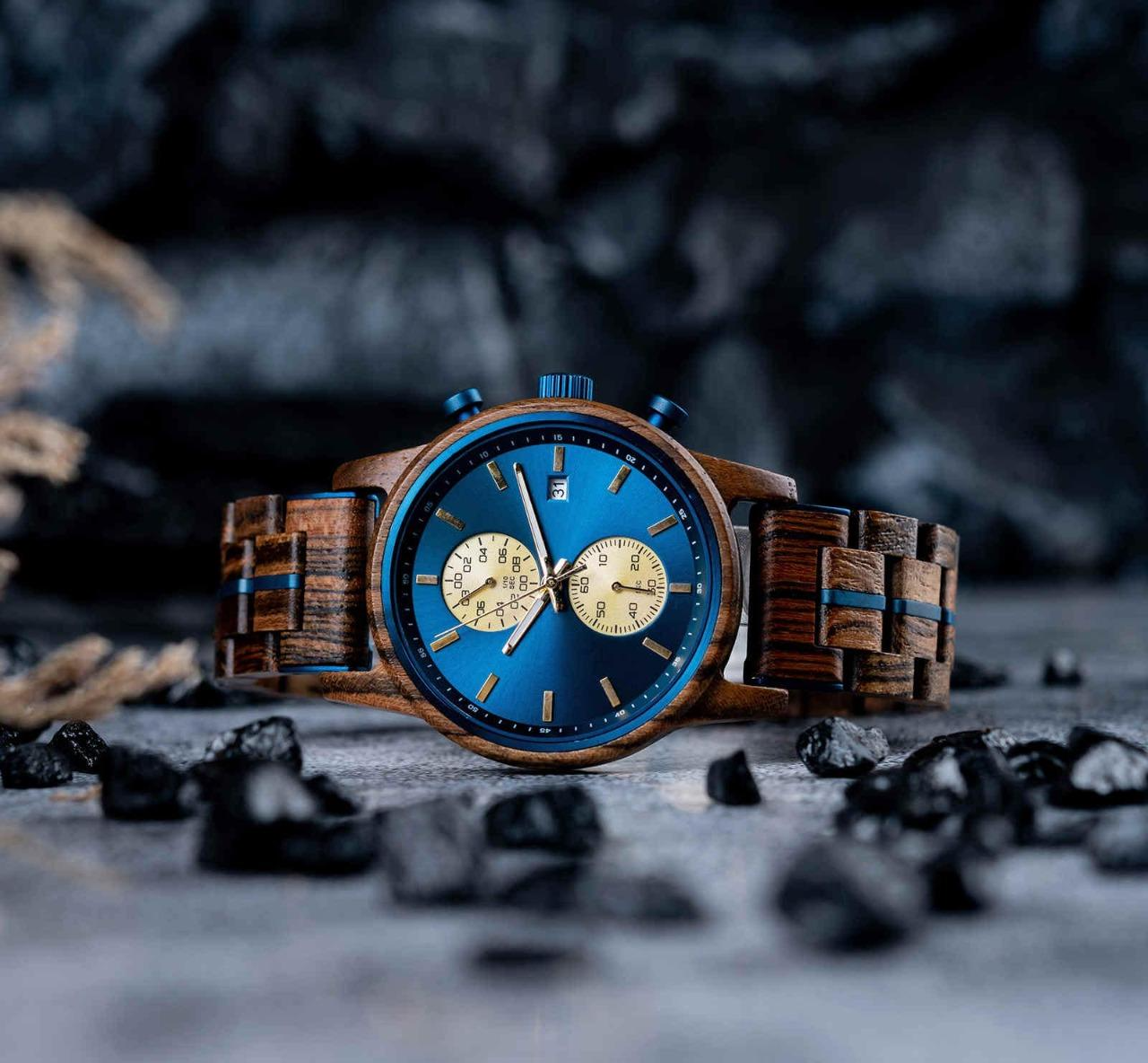 A front view of the Celestial Oak men's wooden watch with a blue dial and gold subdials, positioned on a textured dark surface and surrounded by small dark rocks.