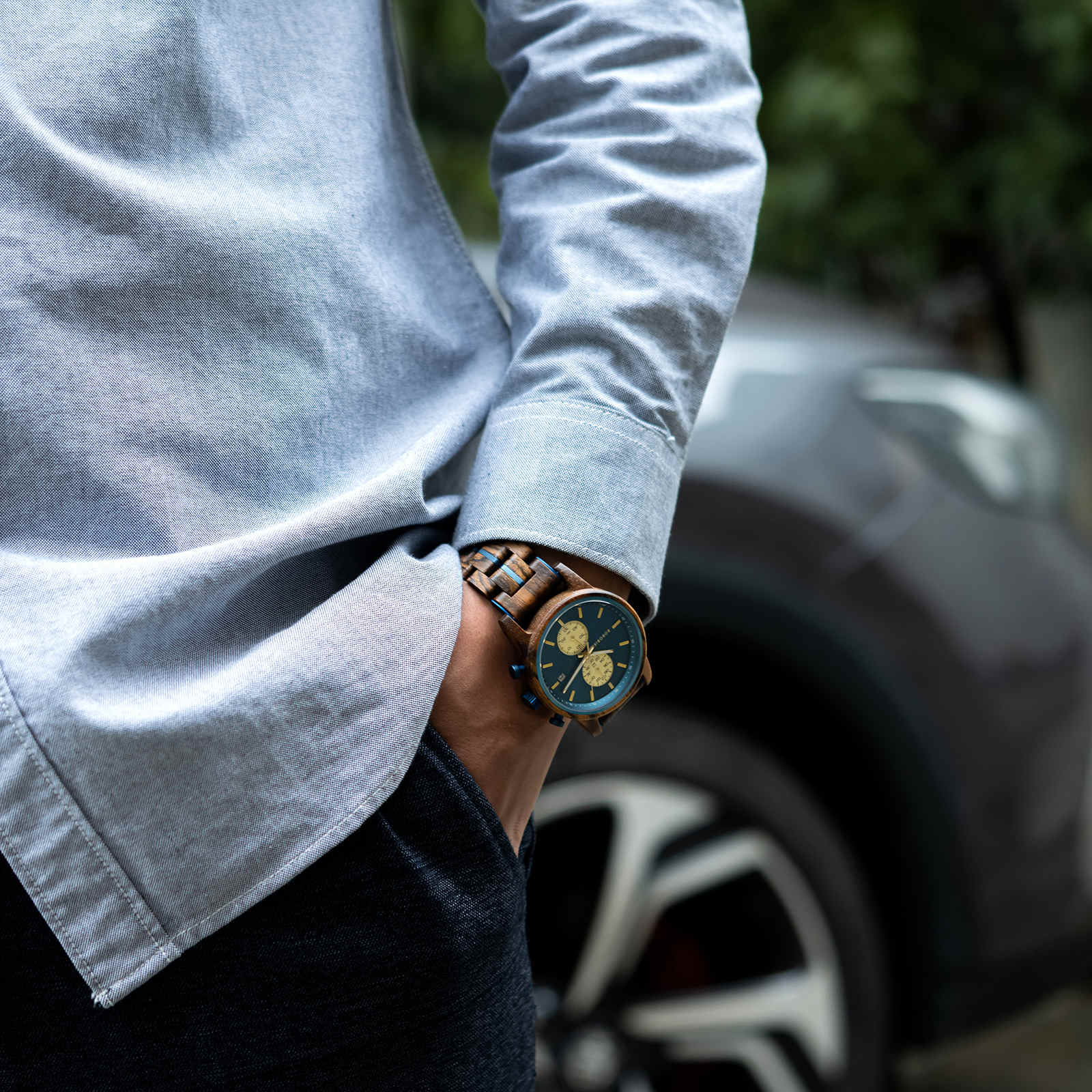 A man wearing the Celestial Oak men's wooden watch with a blue dial and gold subdials on his left wrist, standing casually with a car in the blurred background.