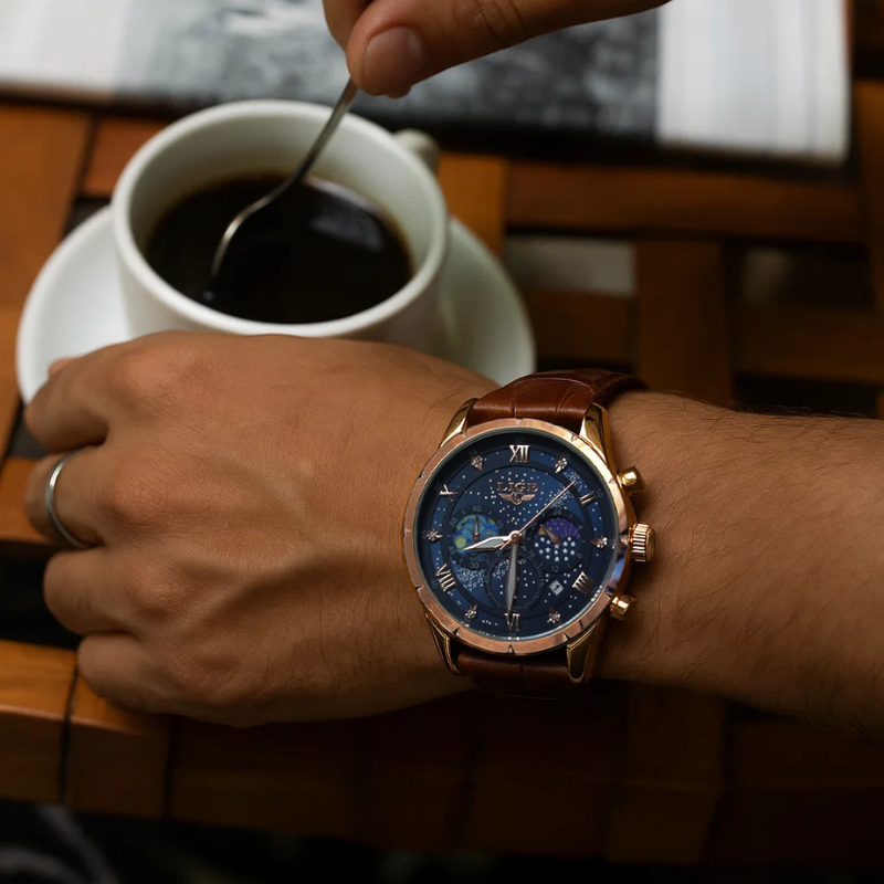 A man's hand wearing the Celestial Timekeeper men's watch with a gold-tone case and brown leather strap, stirring coffee in a white cup.