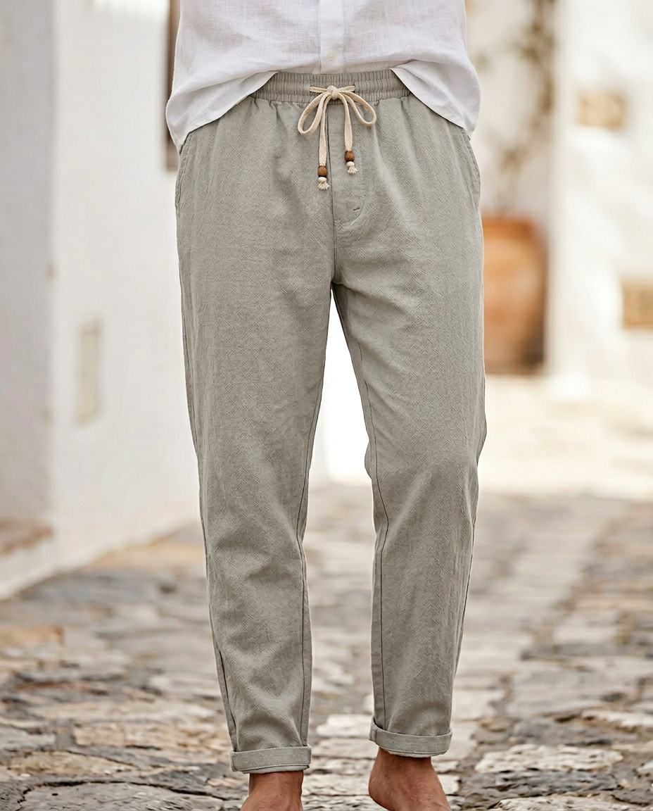 Men's Coastal Breeze Trousers in light gray linen, featuring a drawstring waist and rolled cuffs, styled with a white shirt and shown barefoot on a cobblestone street.