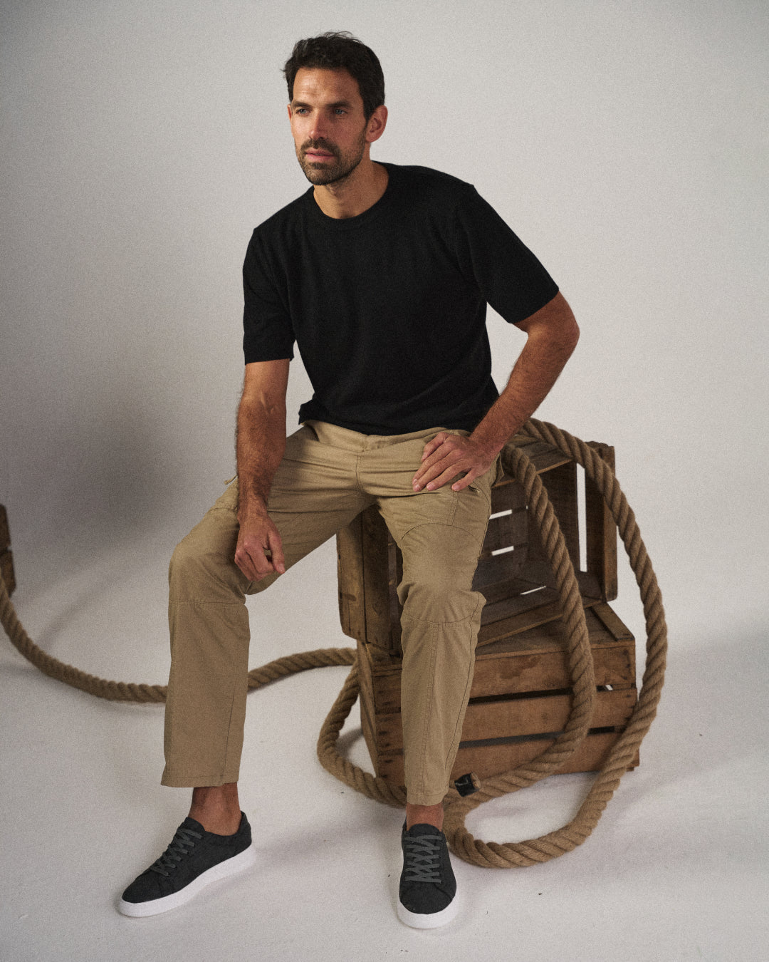 Man sitting, wearing Khaki Coastal Retreat cotton cargo pants with utility pockets, a black t-shirt, and black sneakers, against a white background with wooden crates and rope.