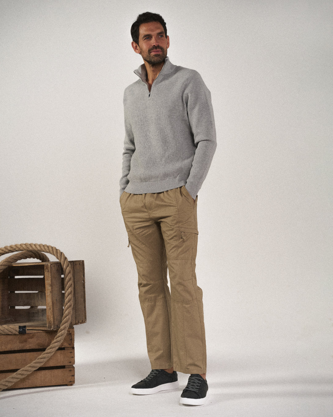Full shot of a man standing, wearing Khaki Coastal Retreat cotton cargo pants with utility pockets, a grey quarter-zip sweater, and black sneakers, posed against a white background with wooden crates and rope.